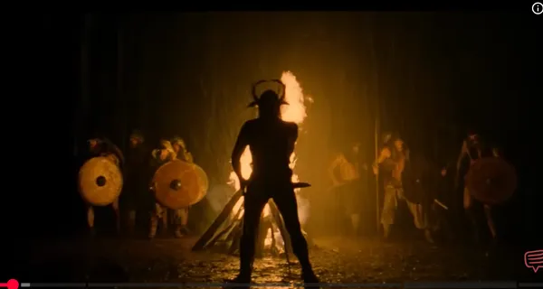 Silhouette of a warrior with horns in front of a fire, surrounded by armed men.