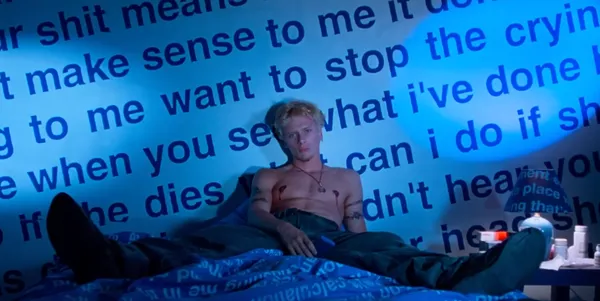 A person sits shirtless on a bed with oversized text on the wall.