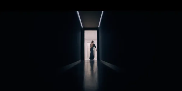 A lone person walks down a dimly lit hallway towards a bright doorway.