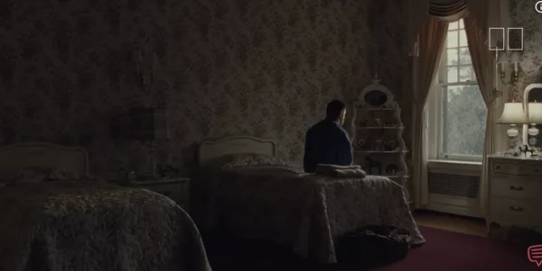 A person sitting on a bed in a dimly lit room, looking out the window.