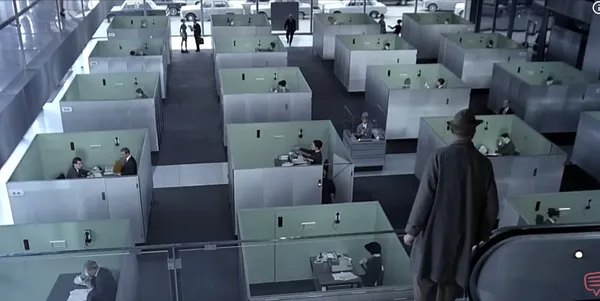 A man descends an escalator overlooking a grid of cubicles in a modern office setting.