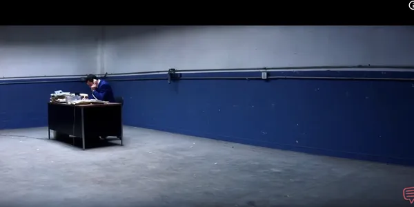 A man is sitting alone at a desk in a large empty room.