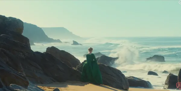 A person in a green dress gazes at the ocean waves crashing on rocky cliffs.