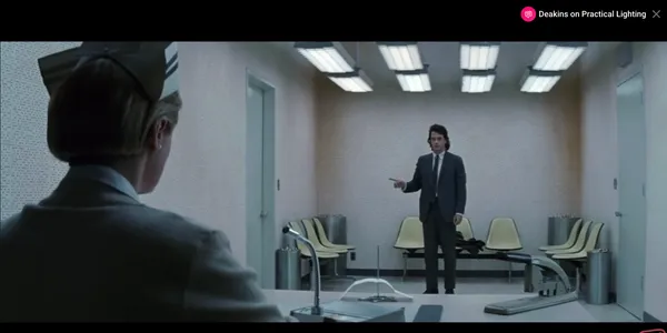 A man in a suit confronts a seated figure in an empty, clinical room.