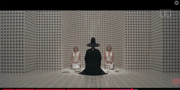 A person in a black robe and hat sits facing two women in a patterned room.