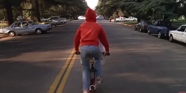 Person riding a bicycle down a road with cars parked on the sides.