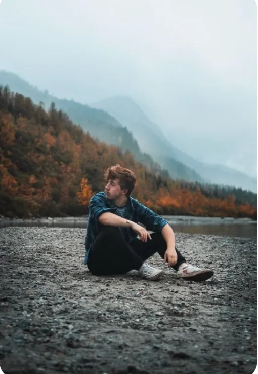 A person seated on a rocky riverbank with misty mountains in the background.