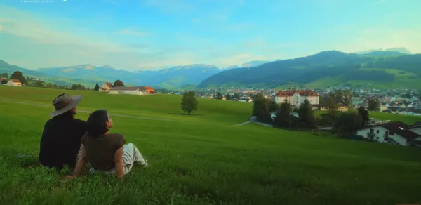 Two people sitting on a grassy hill overlooking a scenic valley and mountains.