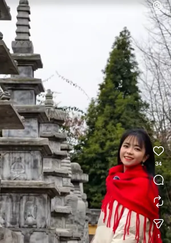 A woman in a red shawl smiling near a stone structure and trees.
